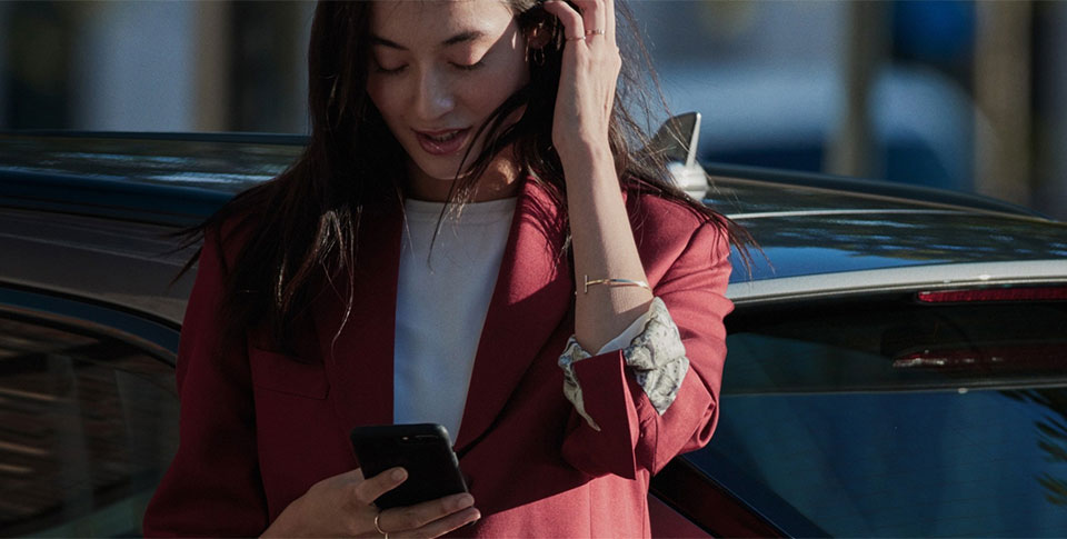 Frau im roten Jackett lehnt an Audi und schaut auf Smartphone runter