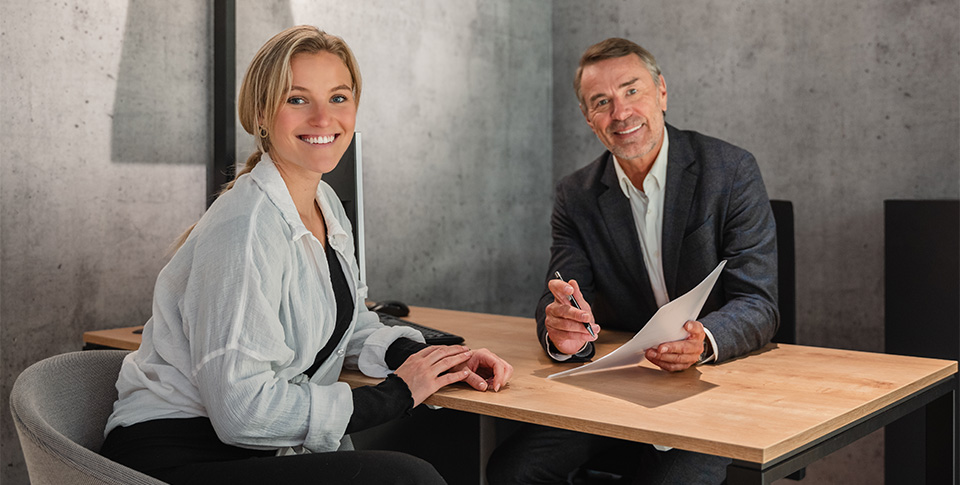 Frau und Mann in Businesskleidung am Schreibtisch. Mann hat eine Mappe und einen Stift in den H&auml;nden.