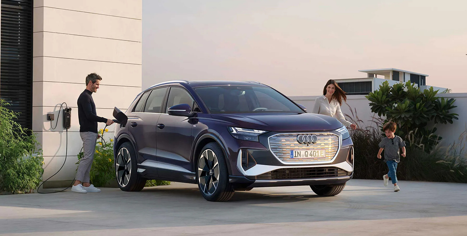 Audi Q4 e-tron in Violett vor Wohngeb&auml;ude. Mann l&auml;dt Fahrzeug. Frau und Kind laufen auf der anderen Seite vorbei